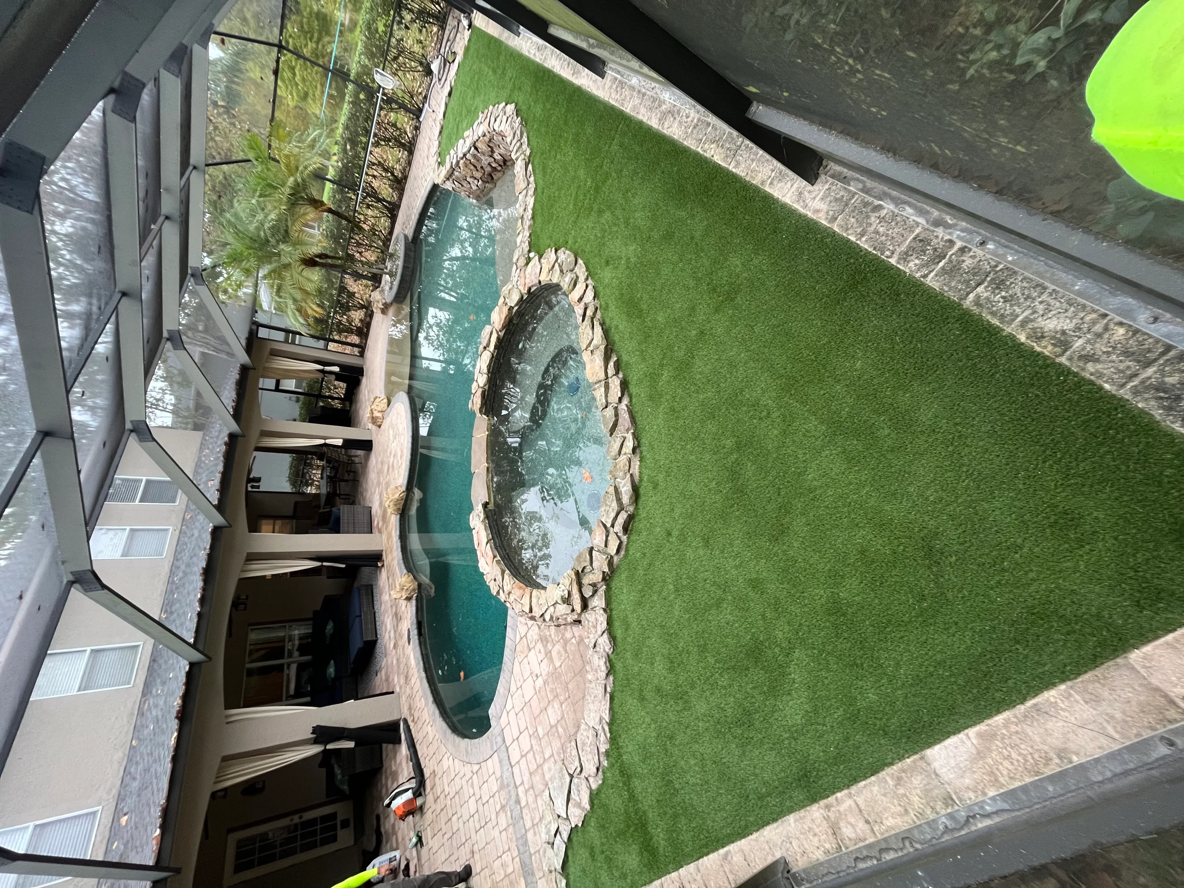 Synthetic turf installation around in-ground pool with stone coping and landscape plantings