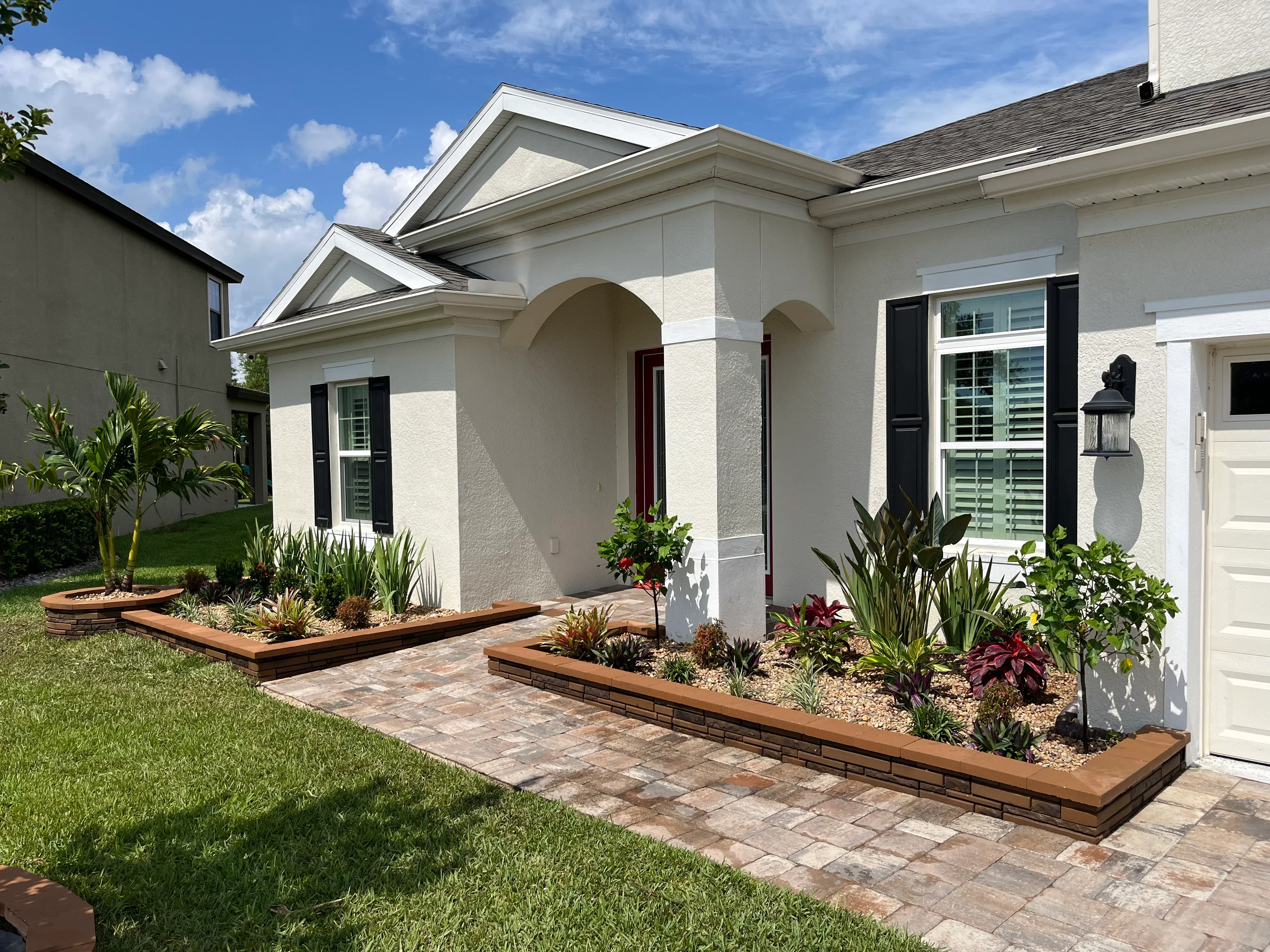 Raised wood planting beds with tropical plants, paver walkway, and manicured lawn in front of white home