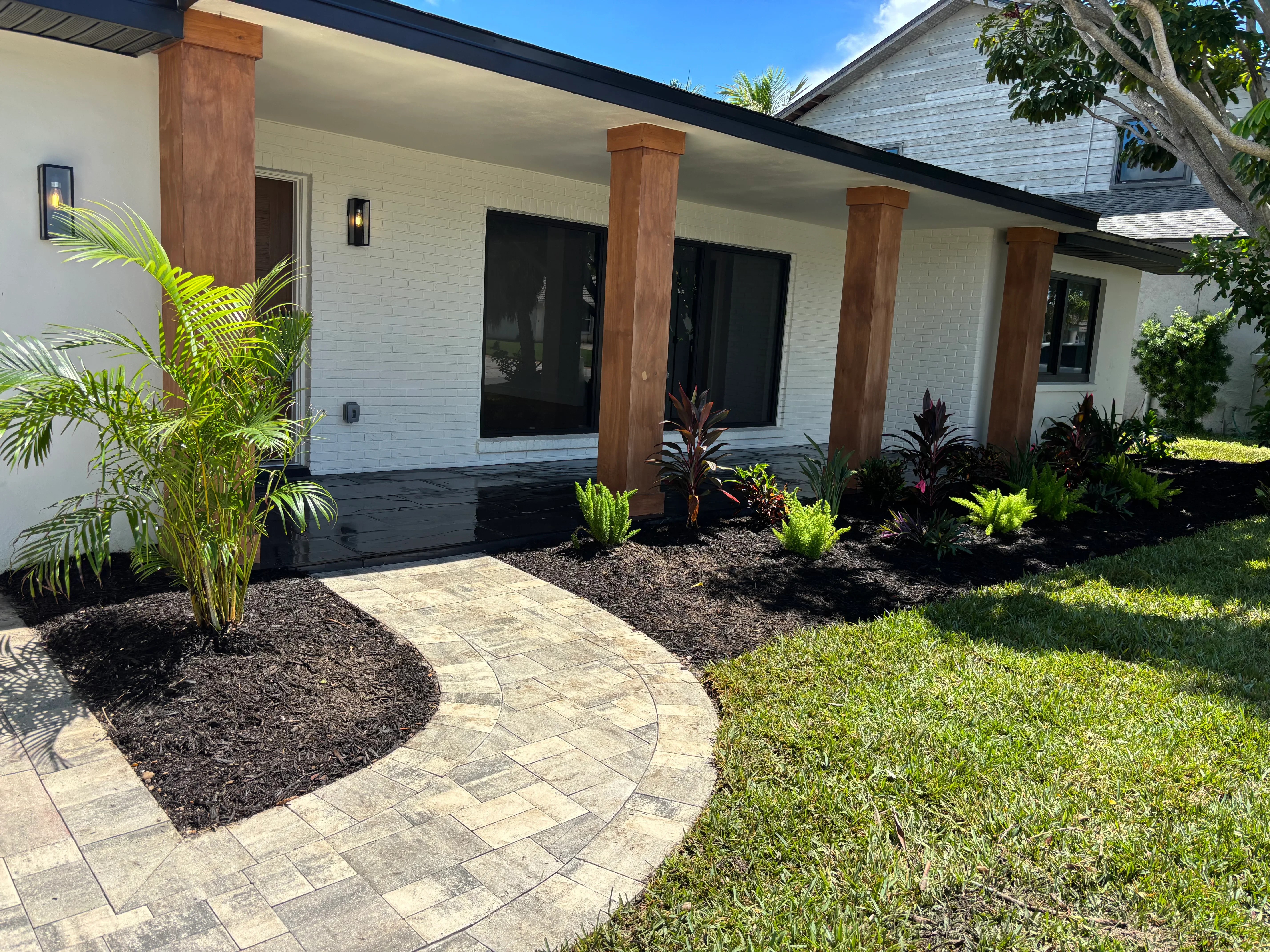 Curved paver walkway with mulch beds, tropical plants, and modern porch with wood posts