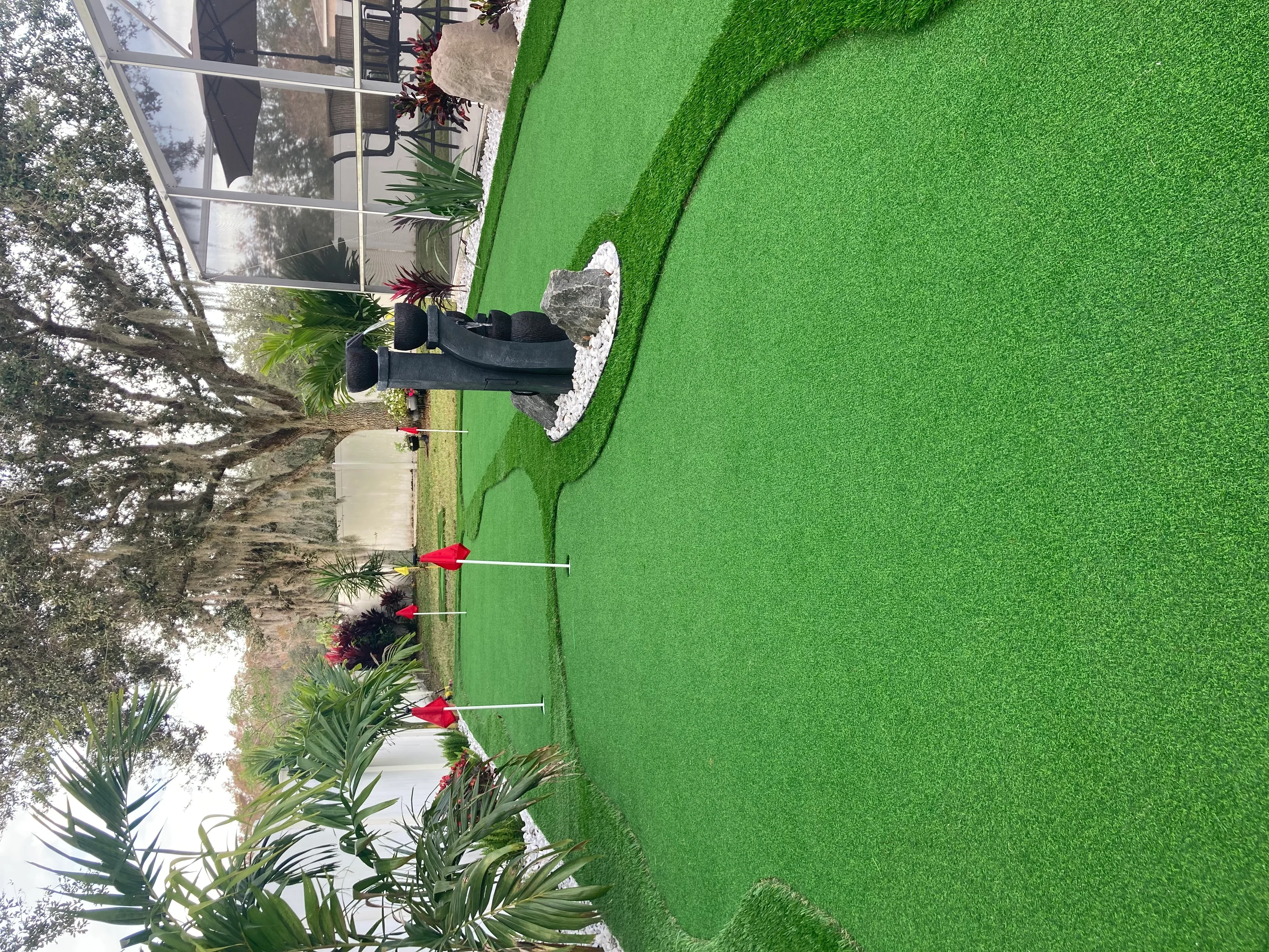 Bright green synthetic turf yard with tropical plants, ornamental grasses, and red plant markers along curved bed edges