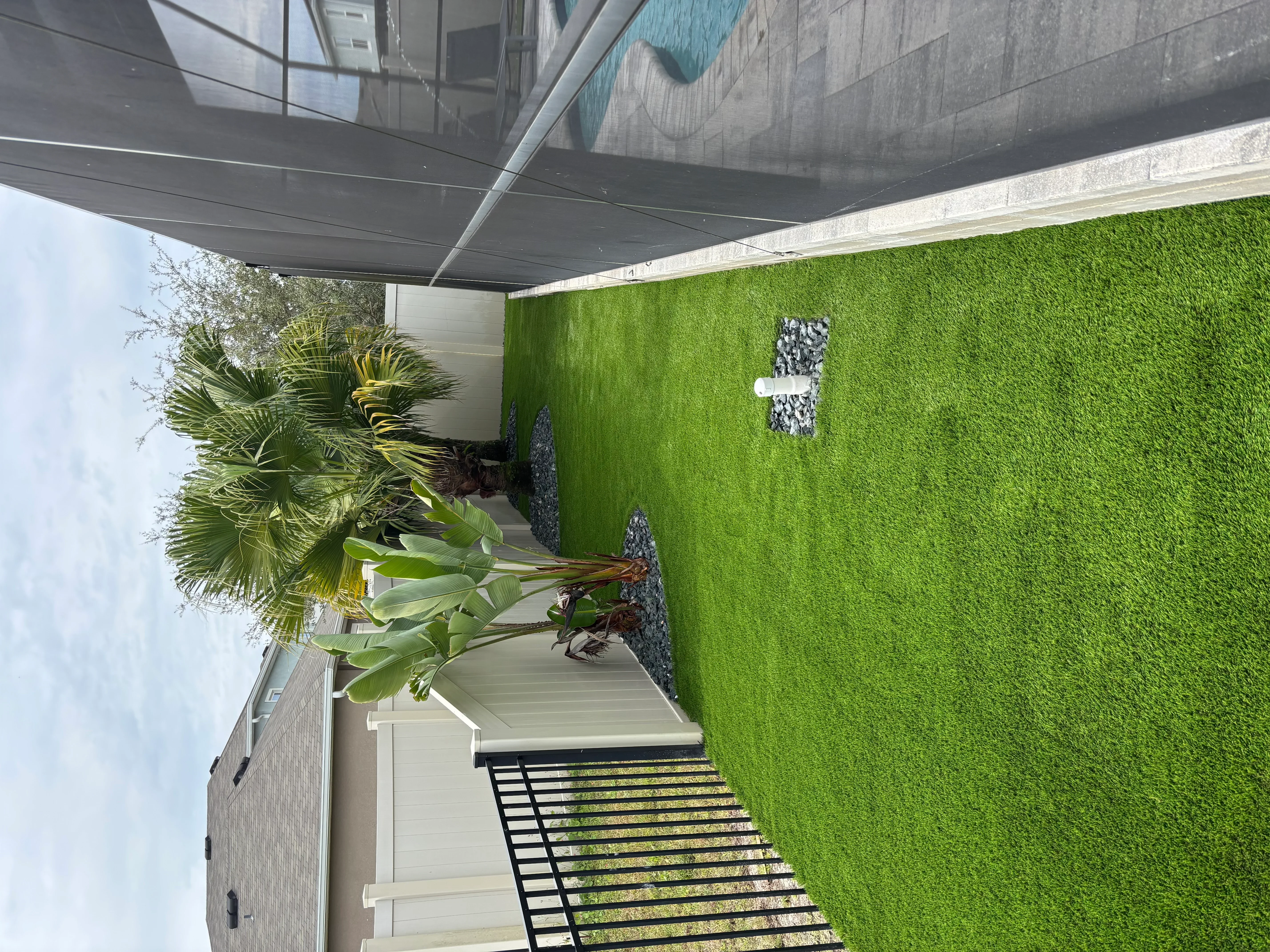 Artificial turf yard with composite deck, retractable shade, tropical plants, and stone accent wall