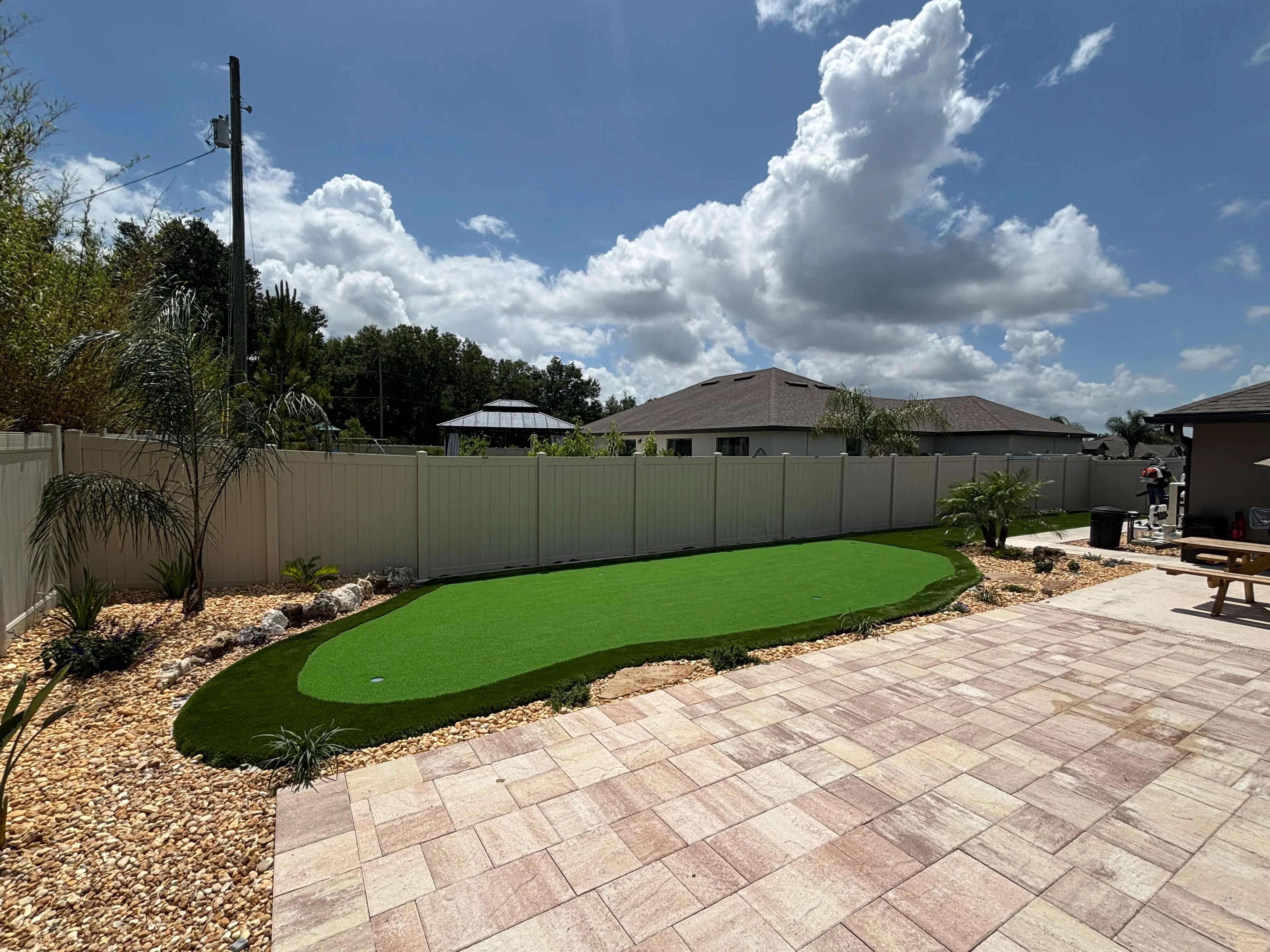 Artificial putting green with paver patio, gravel borders, tropical plants, and composite fencing