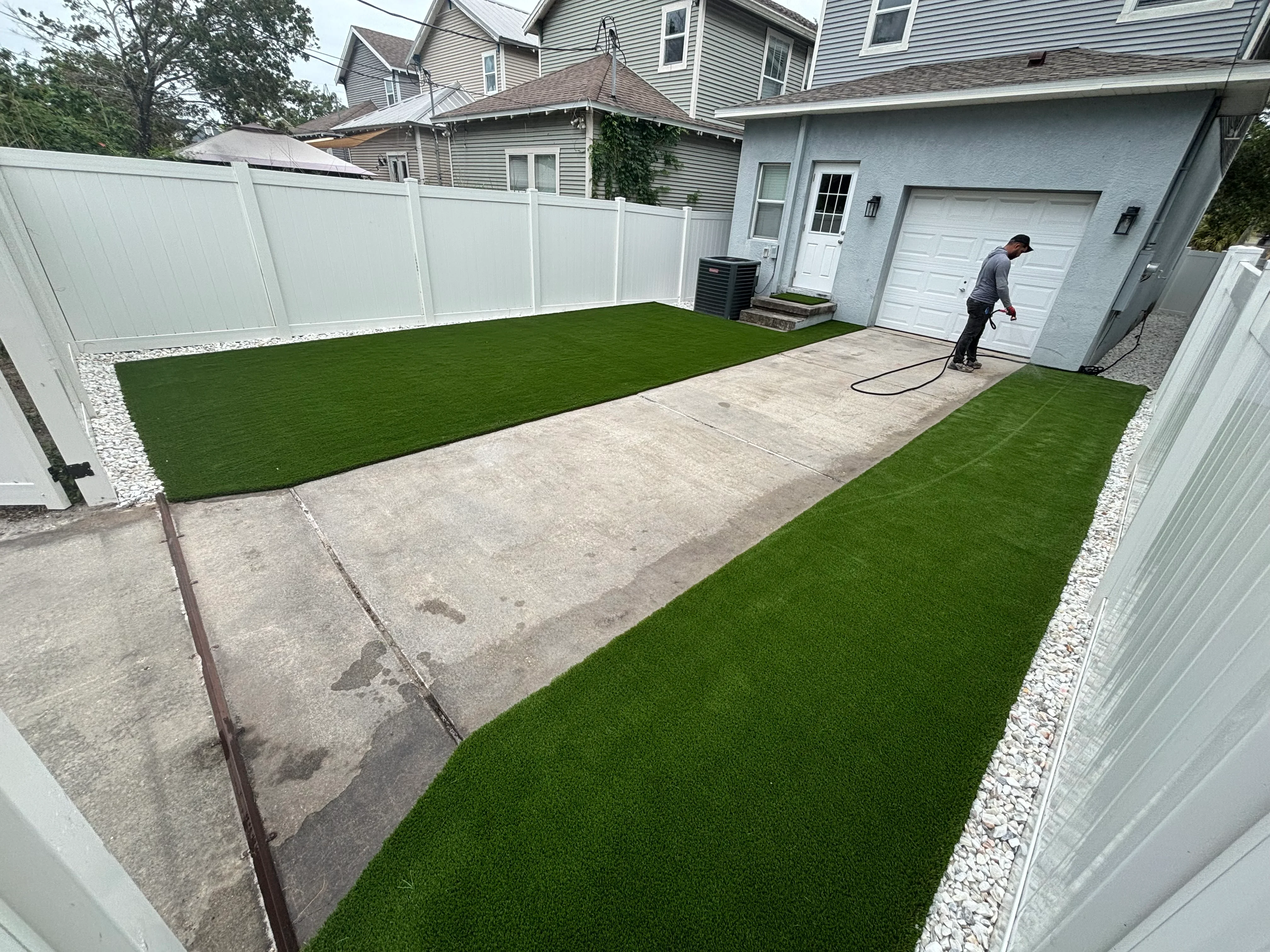 Backyard with synthetic turf, concrete patio, white vinyl fence, and river rock edging