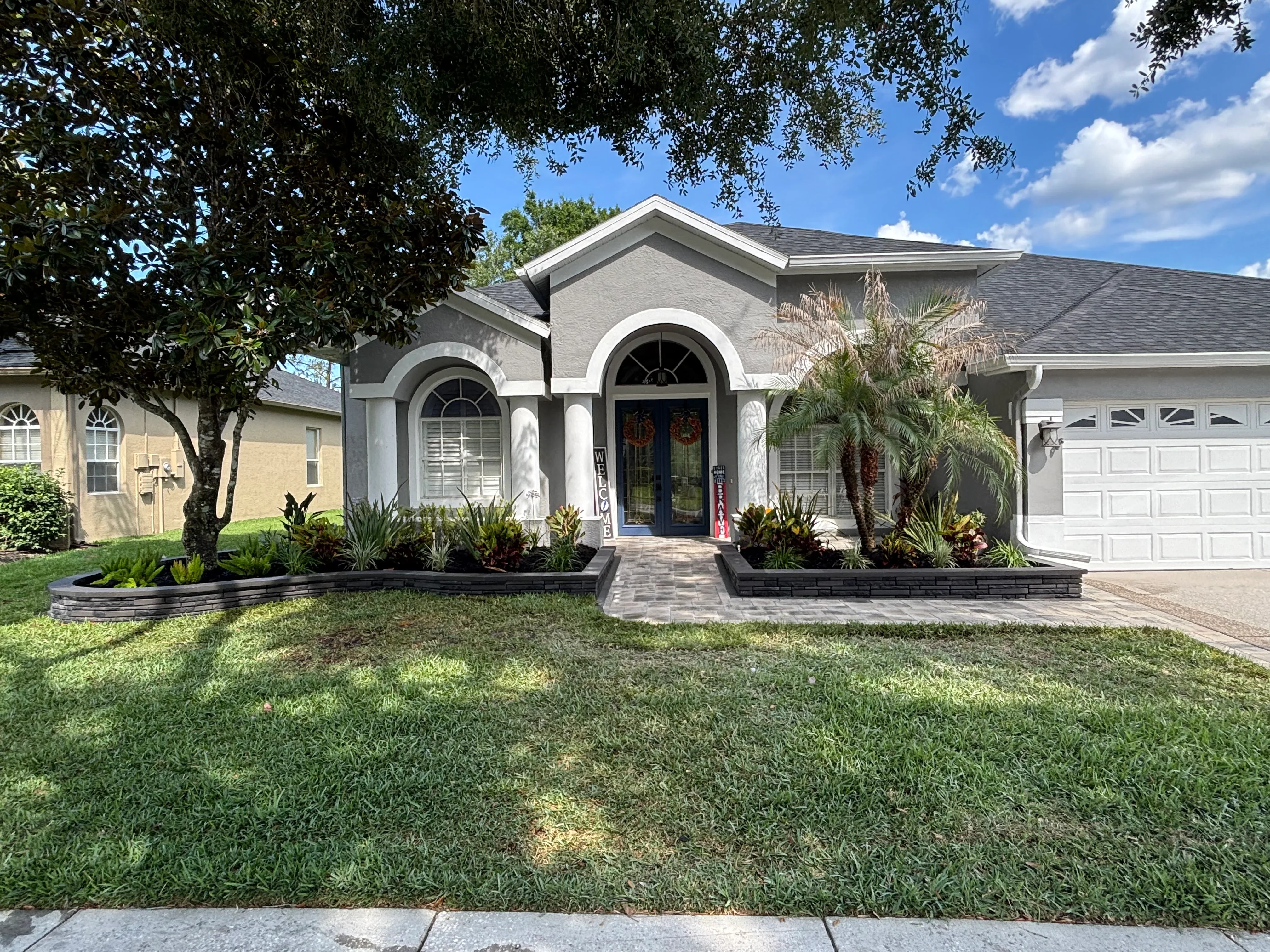 Front yard with raised black-edged planting beds, tropical plants, palms, and paver walkway