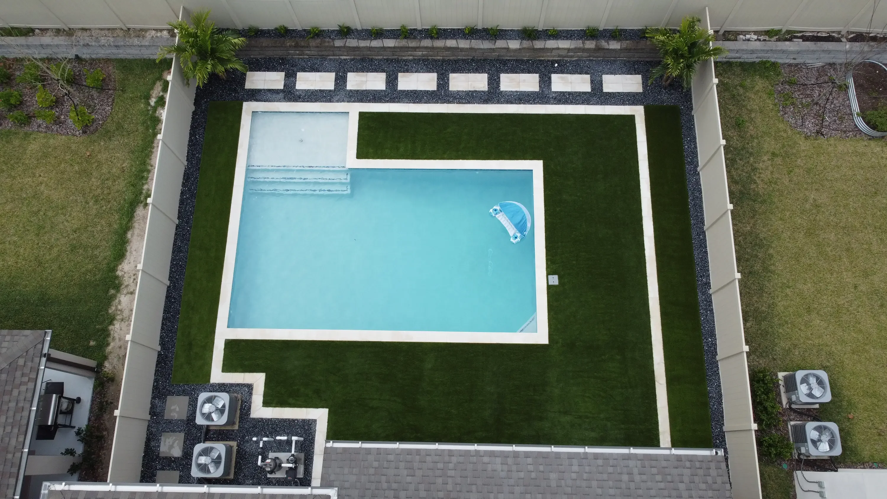 Aerial view of rectangular pool with synthetic turf, white concrete borders, and dark aggregate landscaping