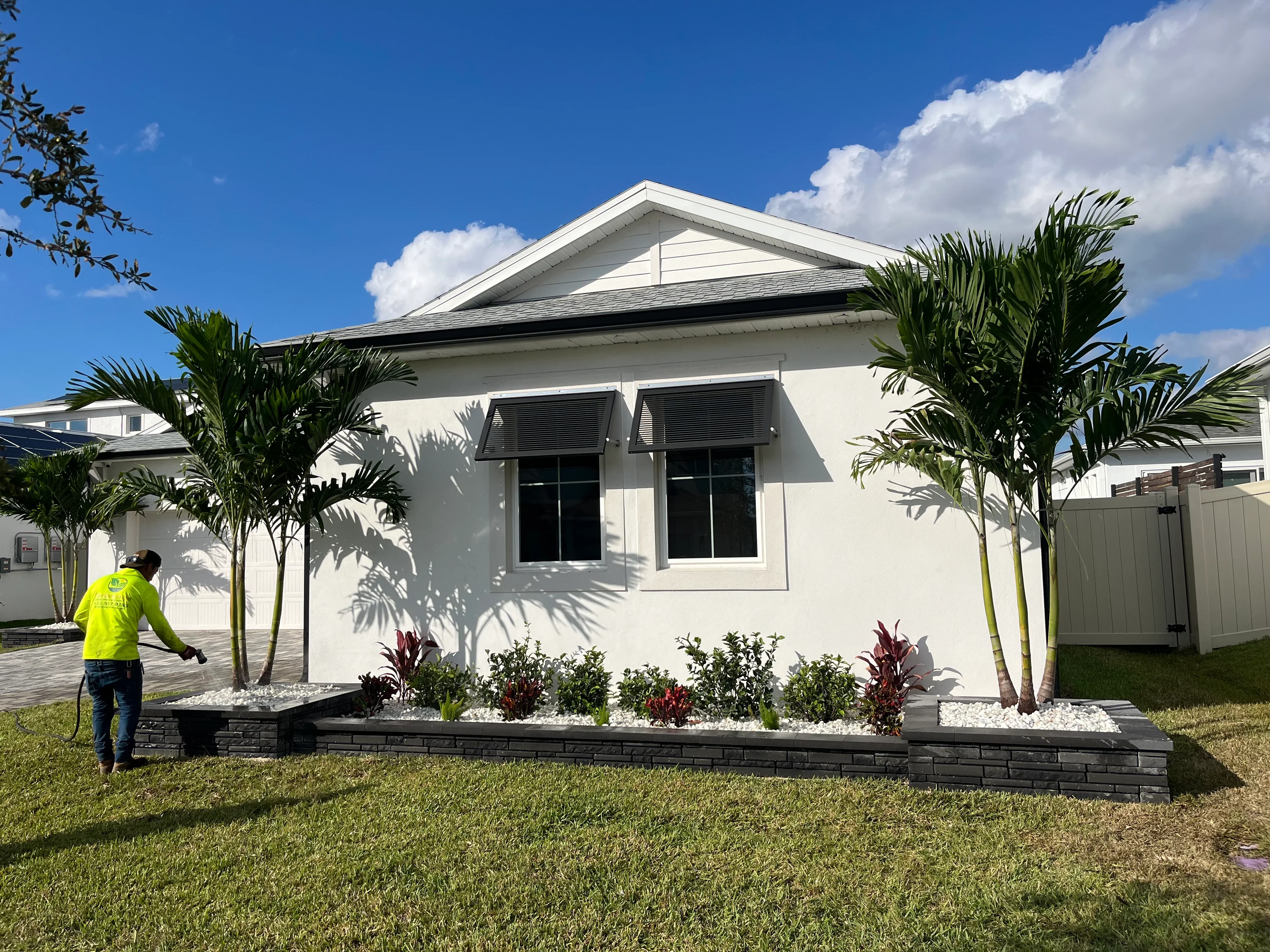 Raised landscape bed with black edging, white rock mulch, tropical plants, and palm trees in front of white home