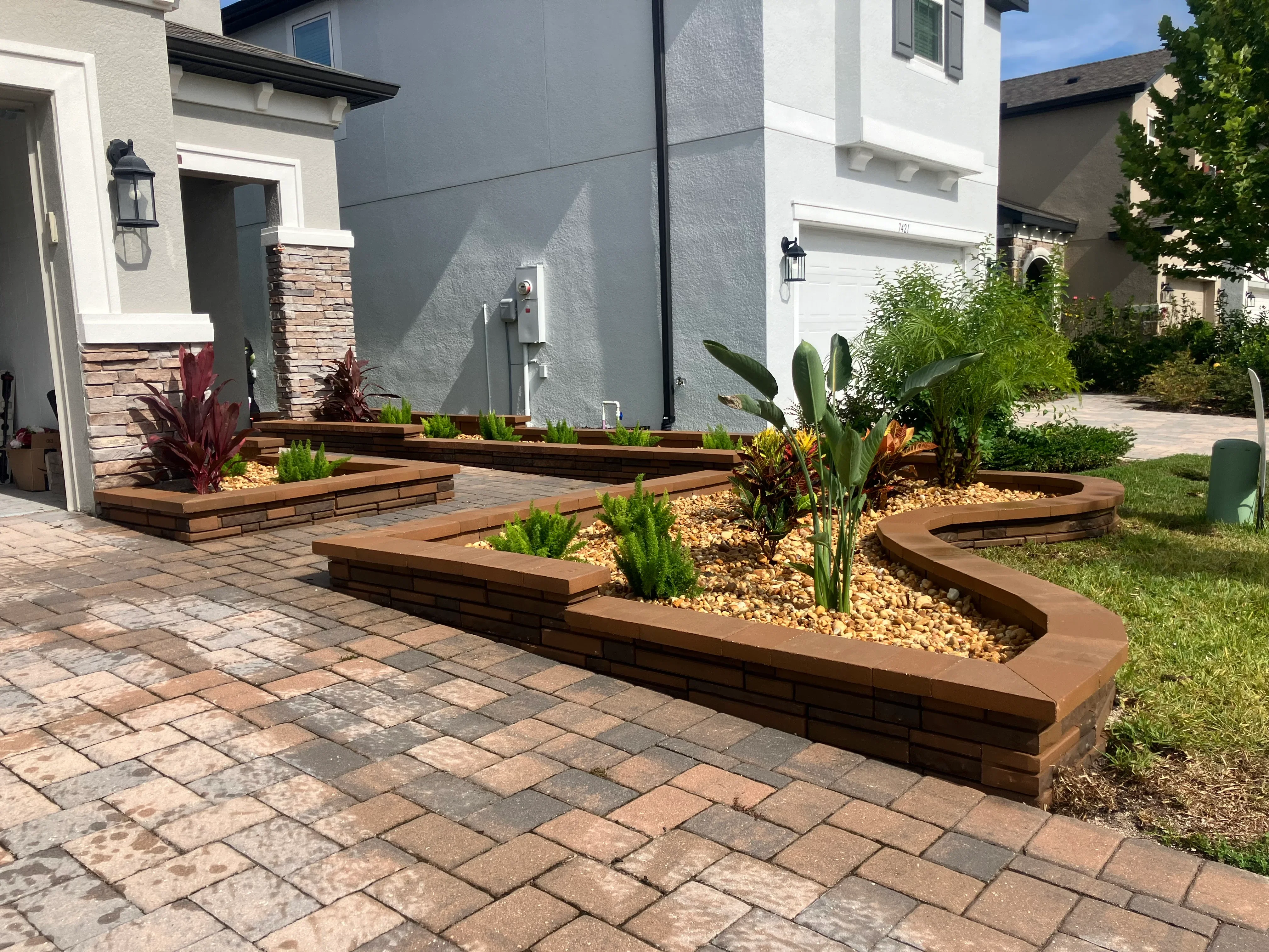 Curved composite wood raised planter beds with gravel fill and tropical plants beside paver patio