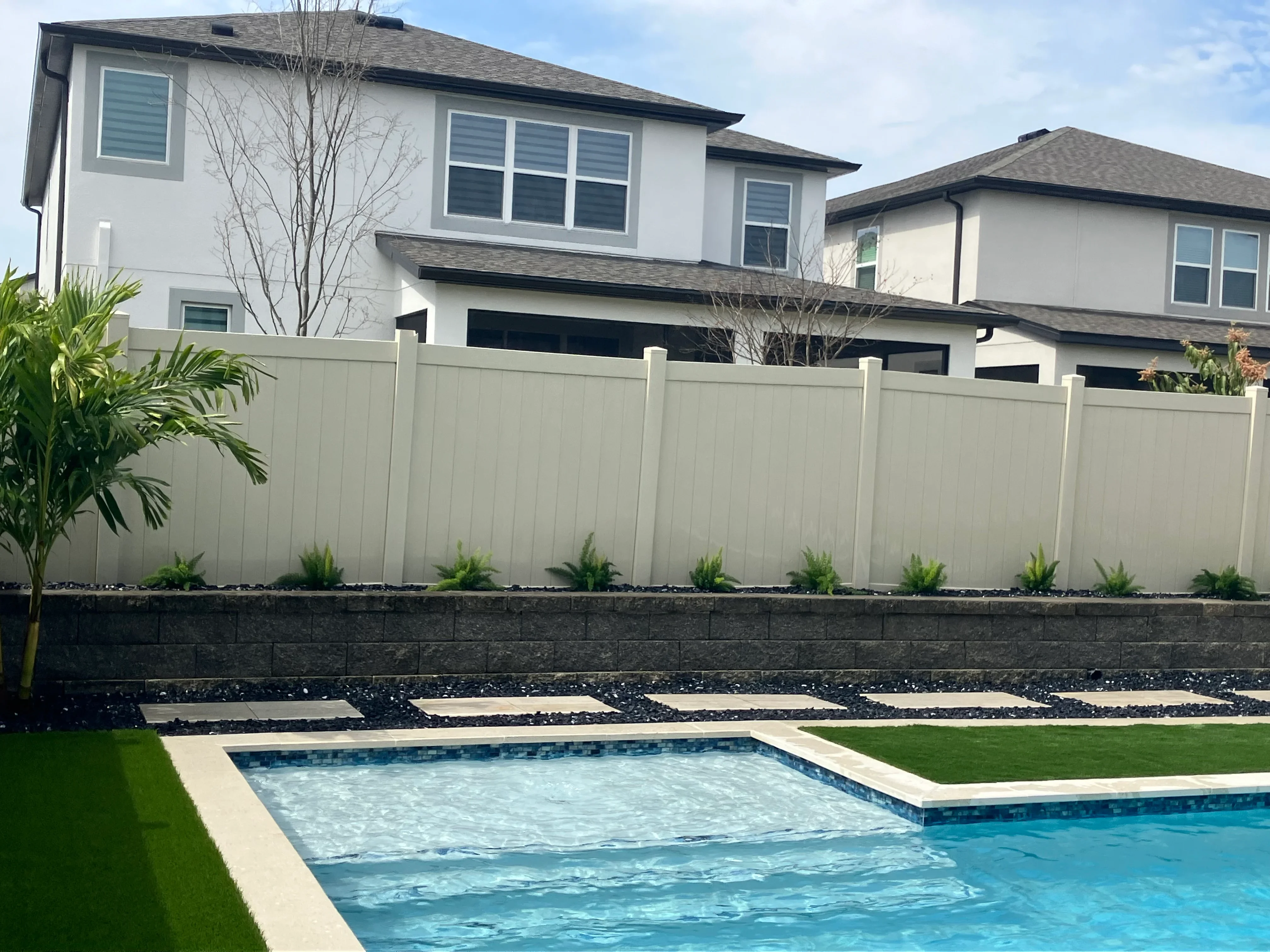 Light gray vinyl privacy fence with dark retaining wall and black rock landscaping along pool deck